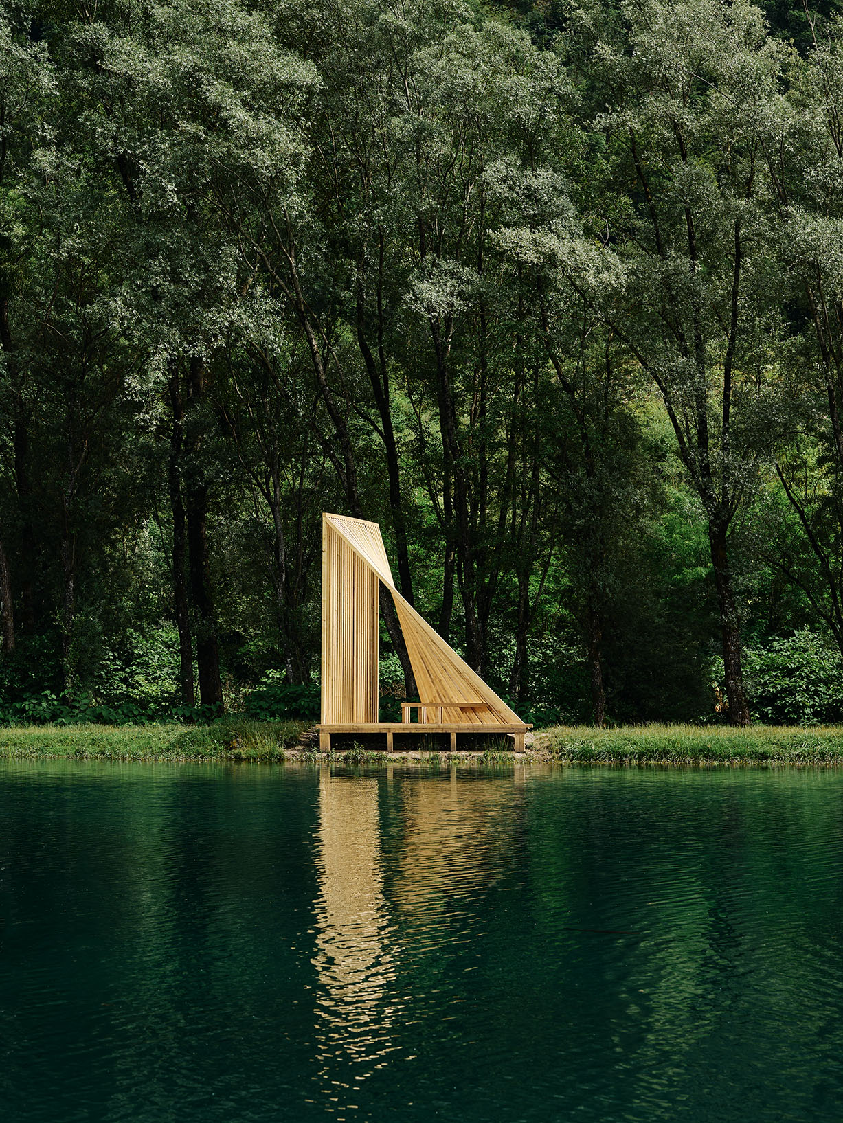 Sculptural timber pavilion composed of vertical slats forming a sail-like silhouette along the lakeshore.